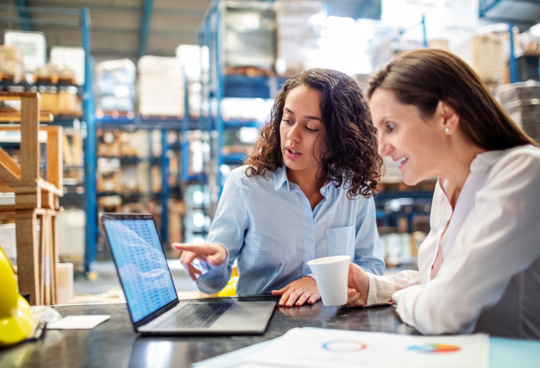 Two women in a warehouse looking at a computer screen, discussing warehouse operations and inventory management.
